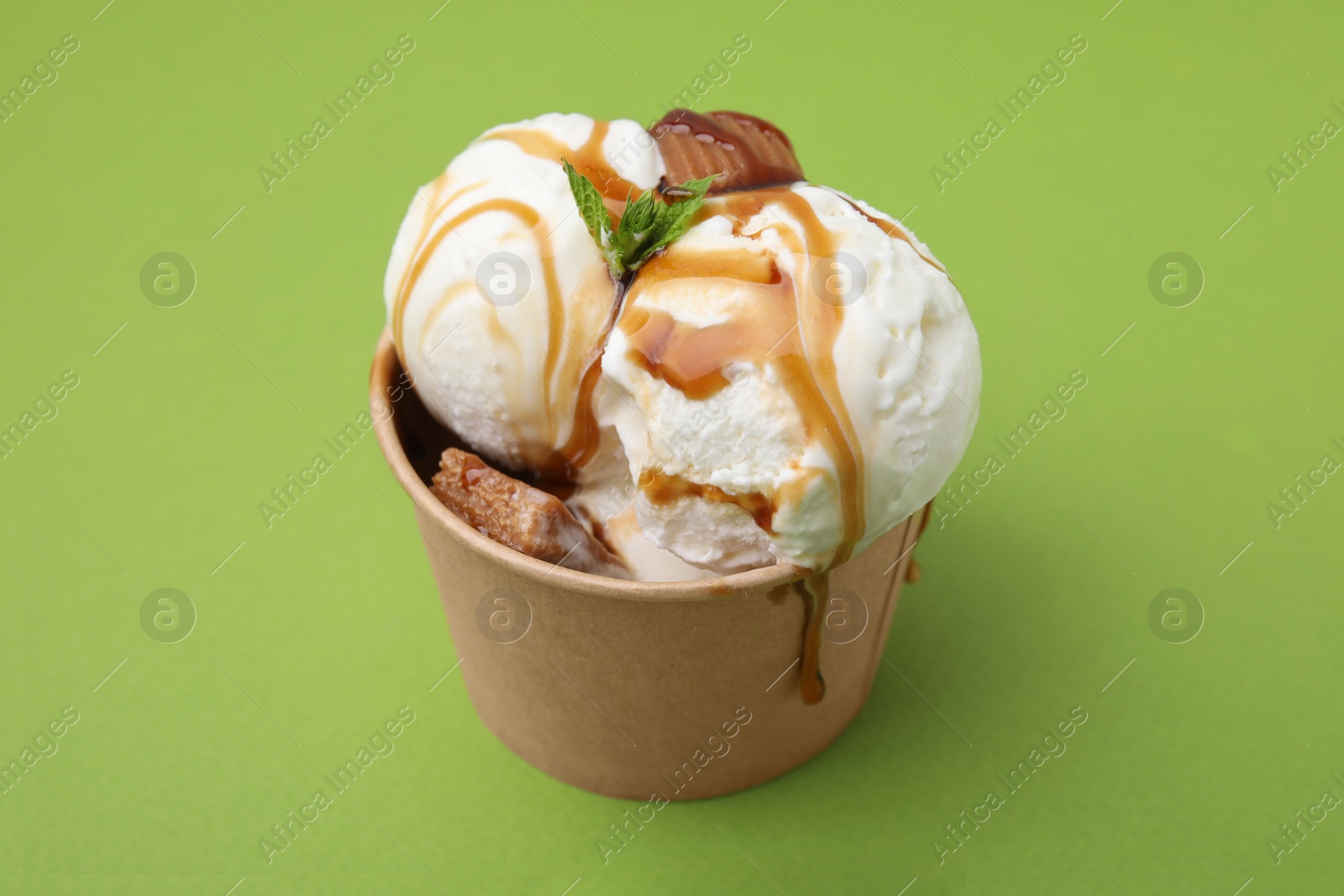 Photo of Scoops of tasty ice cream with mint leaves, caramel sauce and candies in paper cup on green background