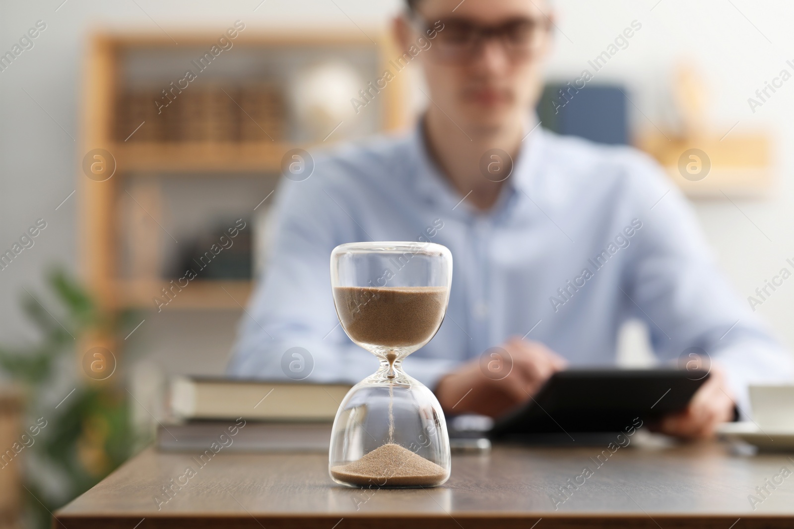 Photo of Hourglass with flowing sand on desk. Man using calculator indoors, selective focus