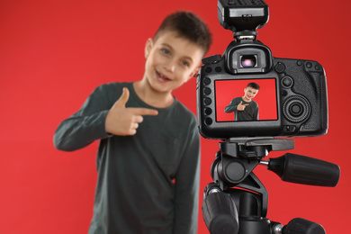 Photo of Cute little blogger recording video against red background, focus on camera