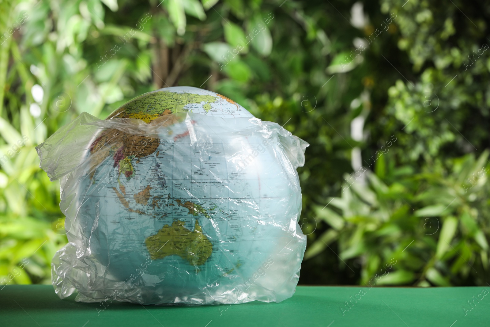 Photo of Globe in plastic bag on table against green leaves, space for text. Environmental conservation