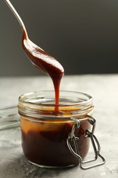 Photo of Taking tasty barbeque sauce from jar with spoon at grey textured table, closeup