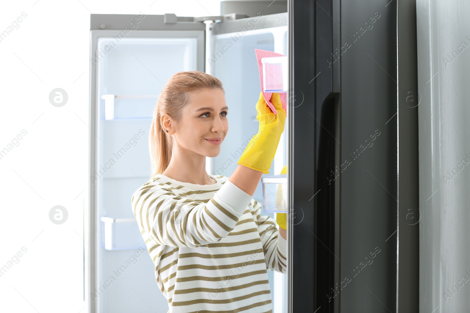 Photo of Woman in rubber gloves cleaning empty refrigerator with rag at home