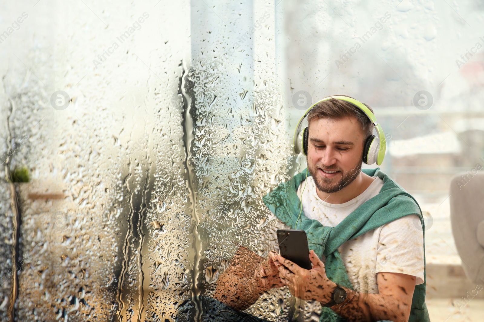 Image of Young man with headphones and mobile device enjoying music at home, view through wet window