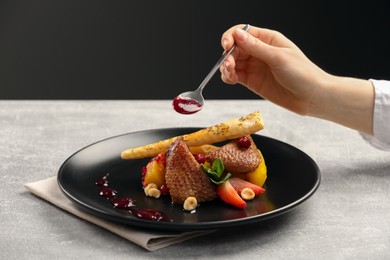 Food stylist adding sauce to delicious dish with chicken, parsnip and strawberries for photoshoot at grey table in studio, closeup