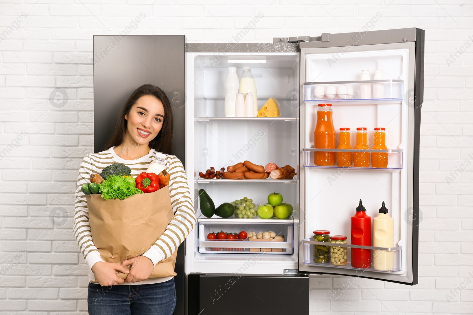 Photo of Young woman with paper bag full of products near open refrigerator indoors