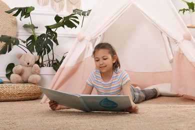 Cute little girl reading book near toy wigwam at home