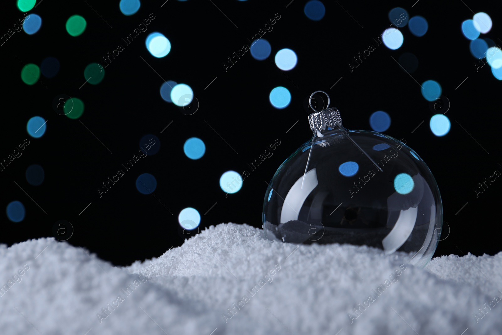 Photo of Transparent Christmas ball on snow against blurred fairy lights, space for text