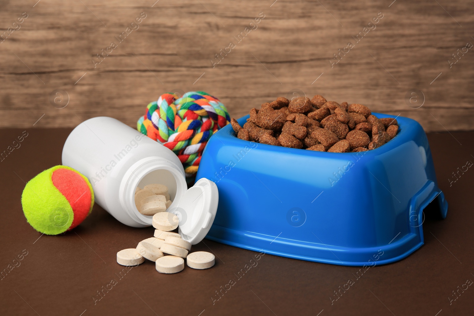 Photo of Bowl with dry pet food, bottle of vitamins and toys on brown surface