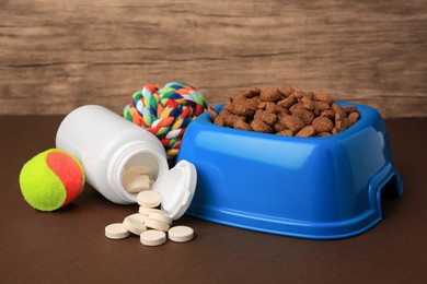 Photo of Bowl with dry pet food, bottle of vitamins and toys on brown surface