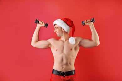 Young muscular man in Santa hat with dumbbells on color background