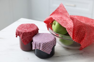 Jars of jams and apples in bowl covered with beeswax food wrap on white table indoors, closeup