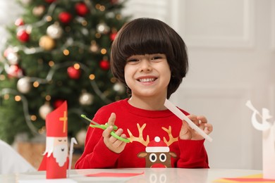Cute little boy cutting paper at table with Saint Nicholas toy indoors