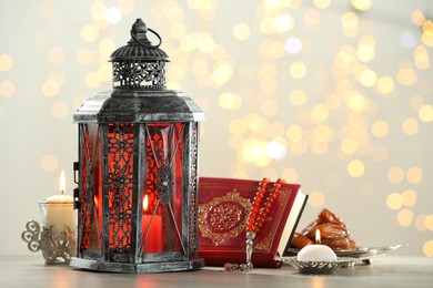 Photo of Arabic lantern, Quran, misbaha, candles and dates on table against blurred lights
