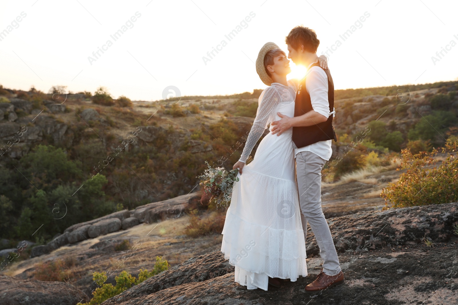 Photo of Happy newlyweds with beautiful field bouquet standing on rock at sunset