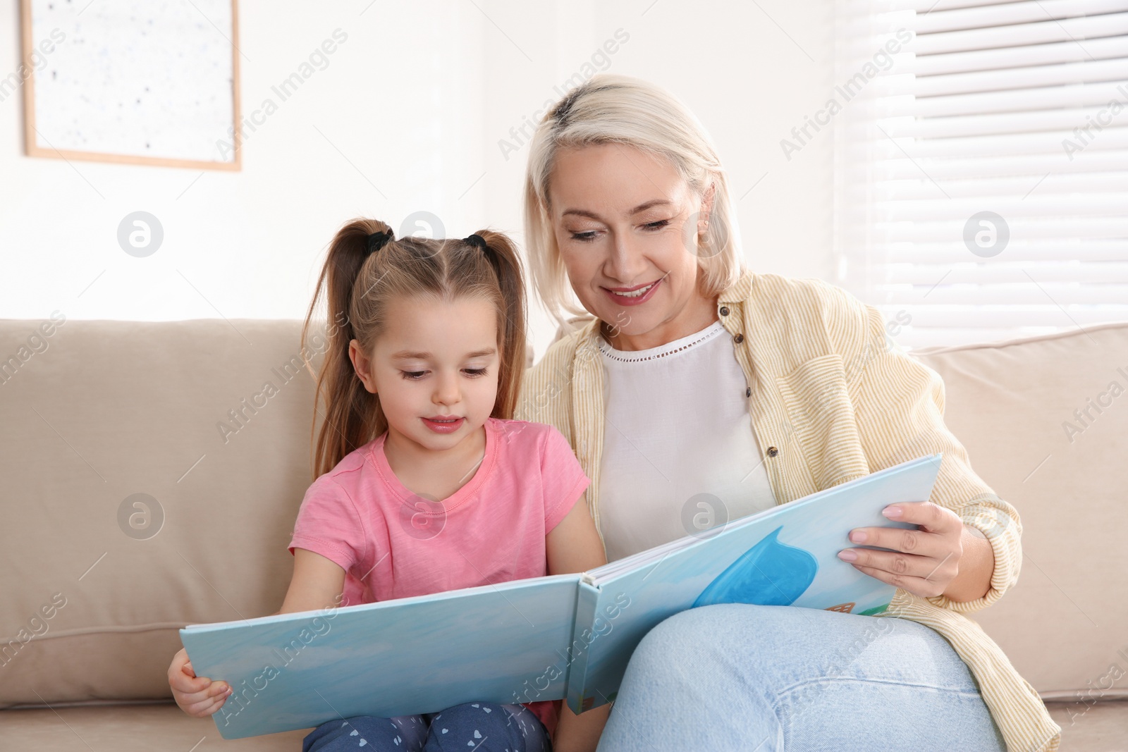 Photo of Mature woman with her little granddaughter reading book together at home