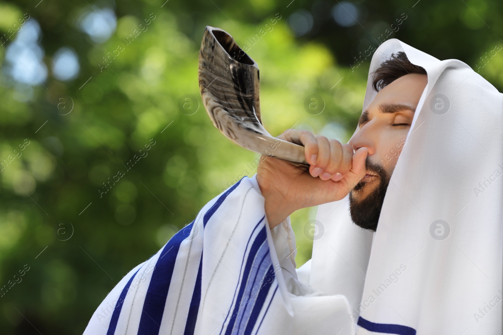 Photo of Jewish man in tallit blowing shofar outdoors. Rosh Hashanah celebration