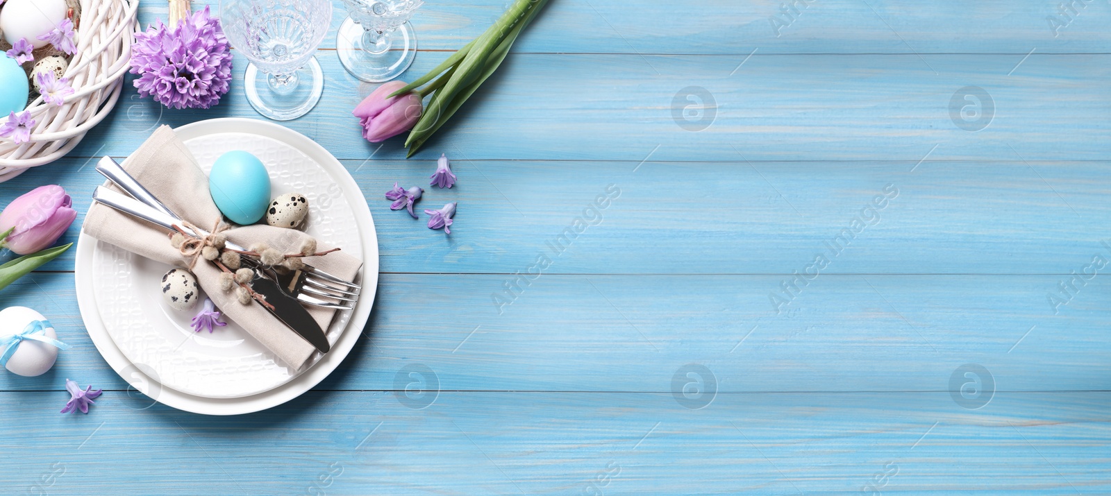 Image of Festive Easter table setting with  painted eggs and floral decor on light blue wooden background, flat lay. Space for text