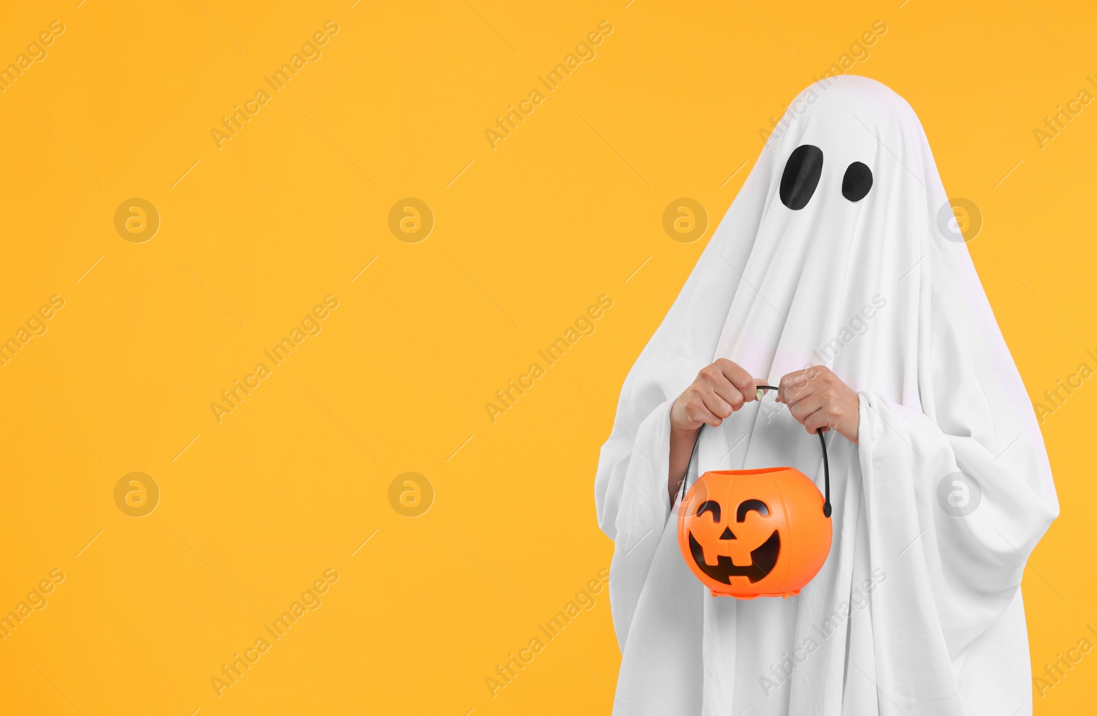 Photo of Woman in white ghost costume holding pumpkin bucket on yellow background, space for text. Halloween celebration