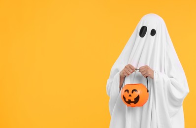 Woman in white ghost costume holding pumpkin bucket on yellow background, space for text. Halloween celebration
