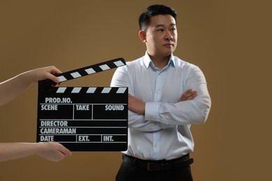 Photo of Asian actor performing while second assistant camera holding clapperboard on brown background, selective focus. Film industry