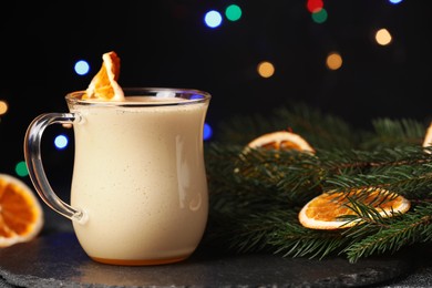 Photo of Glass cup of delicious eggnog with dried orange slice and fir branch on black table