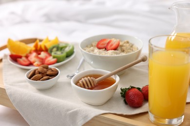 Photo of Tasty breakfast served in bed. Oatmeal, juice, fruits, almonds and honey on wooden tray, closeup