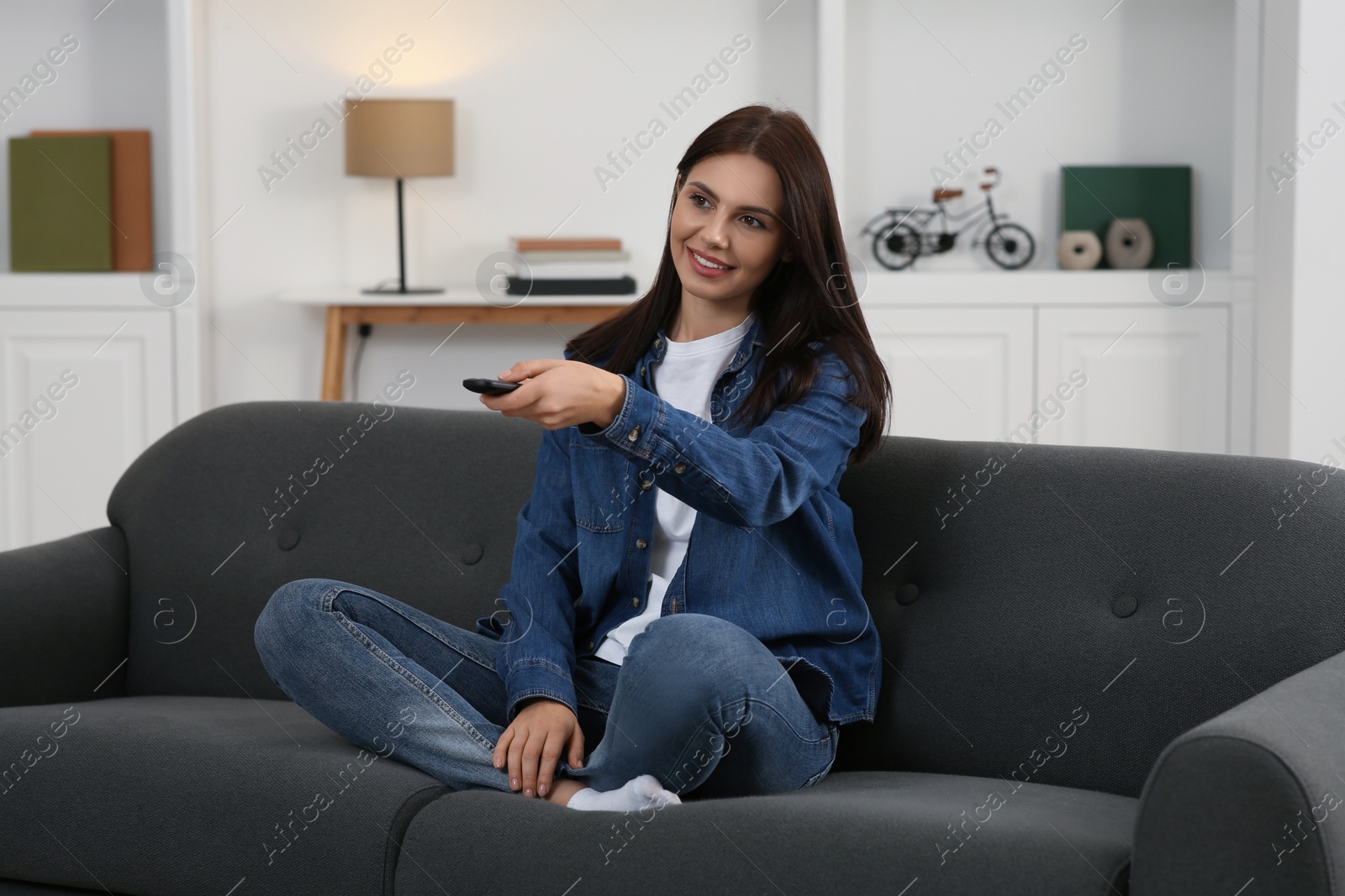 Photo of Happy woman changing TV channels with remote control on sofa at home