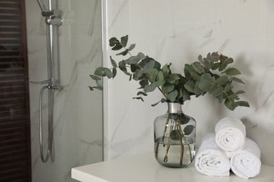 Photo of Rolled towels and glass vase with beautiful eucalyptus branches near shower stall in bathroom. Interior design