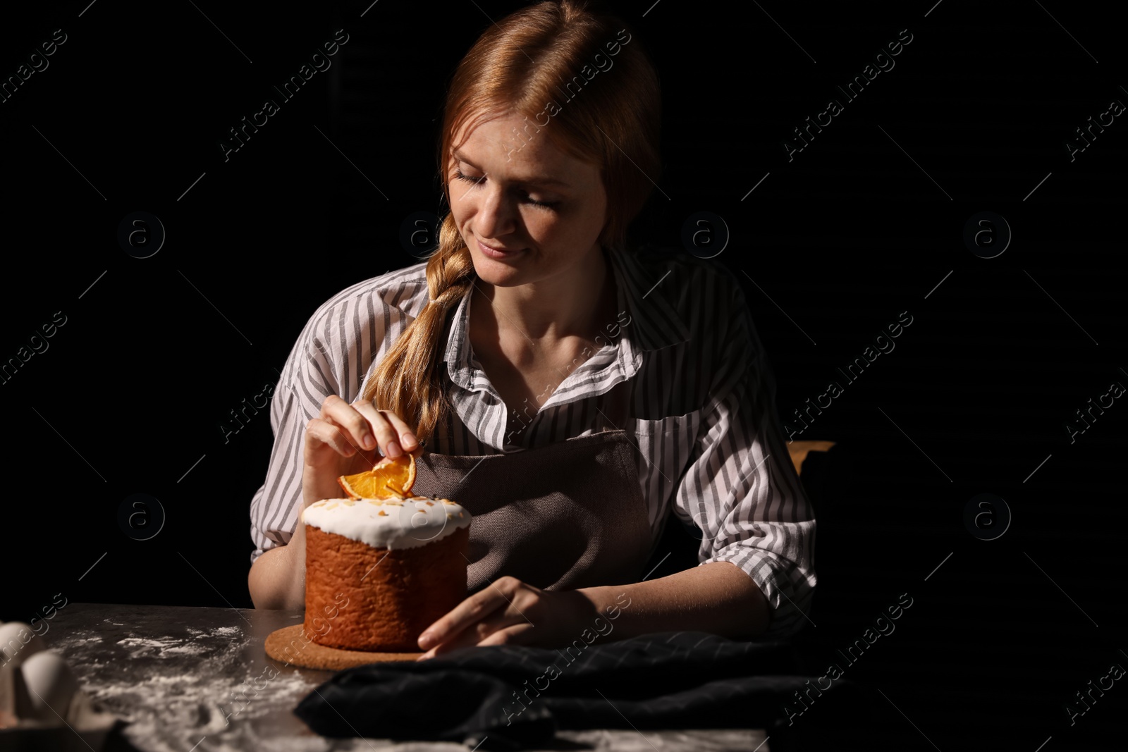 Photo of Young woman decorating traditional Easter cake at table against black background. Space for text