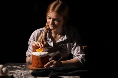 Photo of Young woman decorating traditional Easter cake at table against black background. Space for text