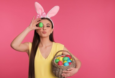 Beautiful woman in bunny ears headband holding basket with Easter eggs on color background, space for text