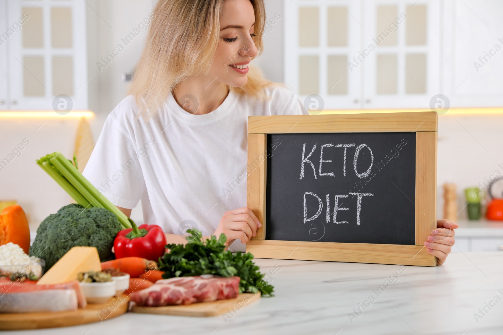 Photo of Woman holding blackboard with phrase Keto Diet at white table in kitchen, closeup