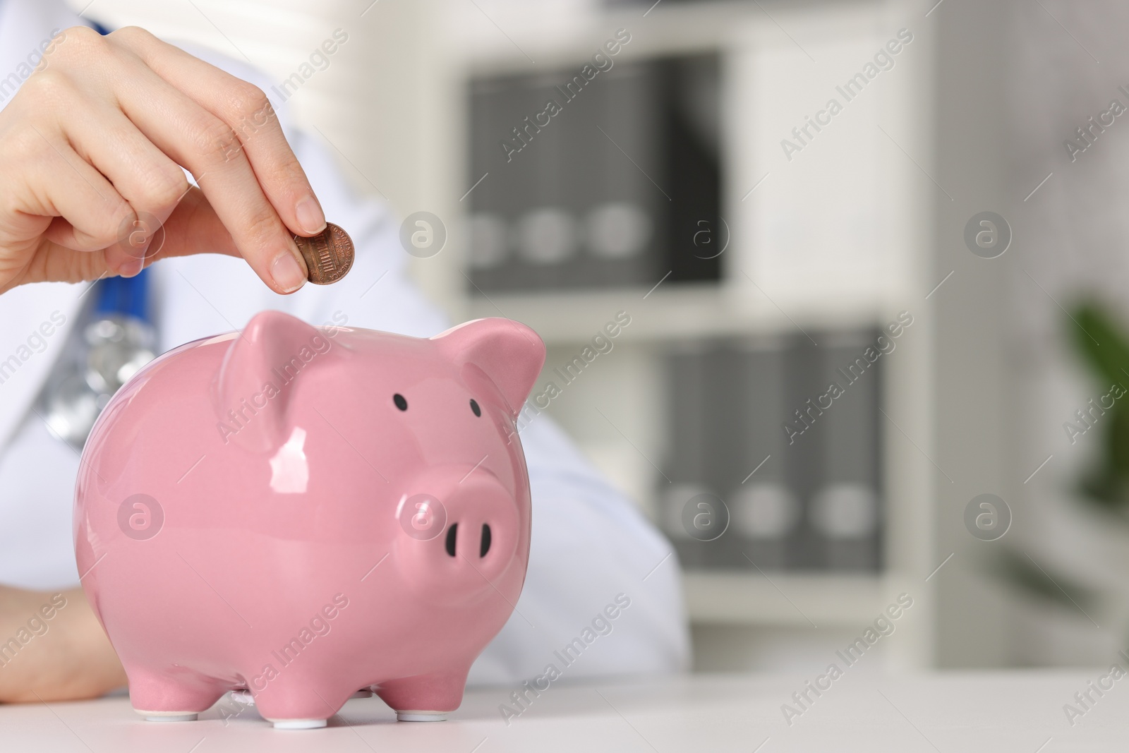 Photo of Doctor putting coin into piggy bank at white table indoors, closeup. Space for text