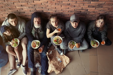 Poor people with plates of food sitting at wall indoors, view from above