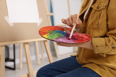 Photo of Woman mixing paints on palette with brush near easel in studio, closeup