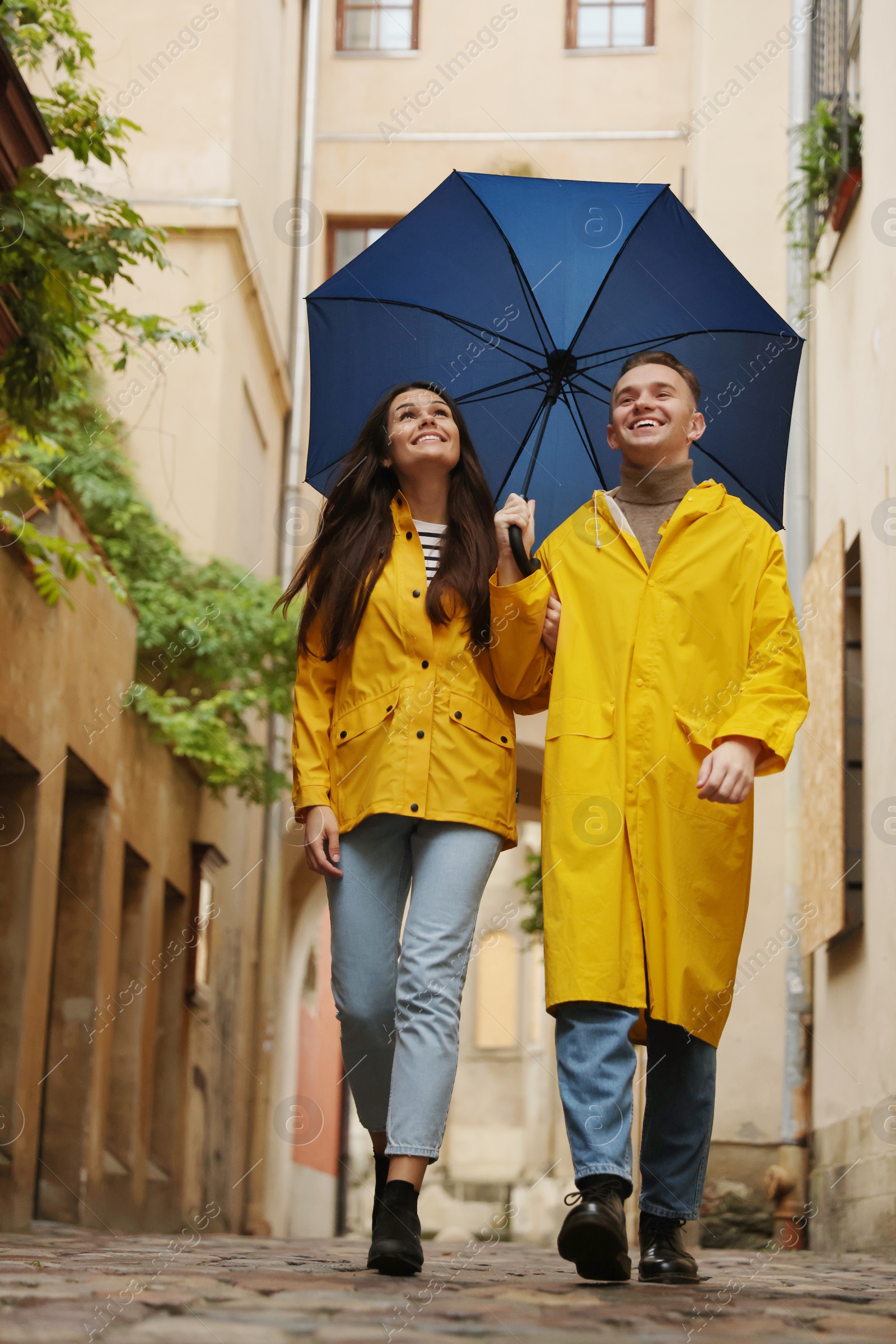 Photo of Lovely young couple with umbrella walking under rain on city street, low angle view