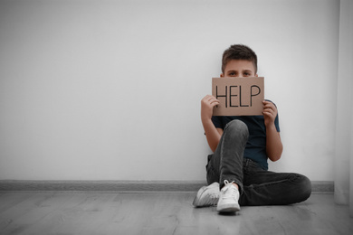 Photo of Sad little boy with sign HELP sitting on floor indoors, space for text. Child in danger