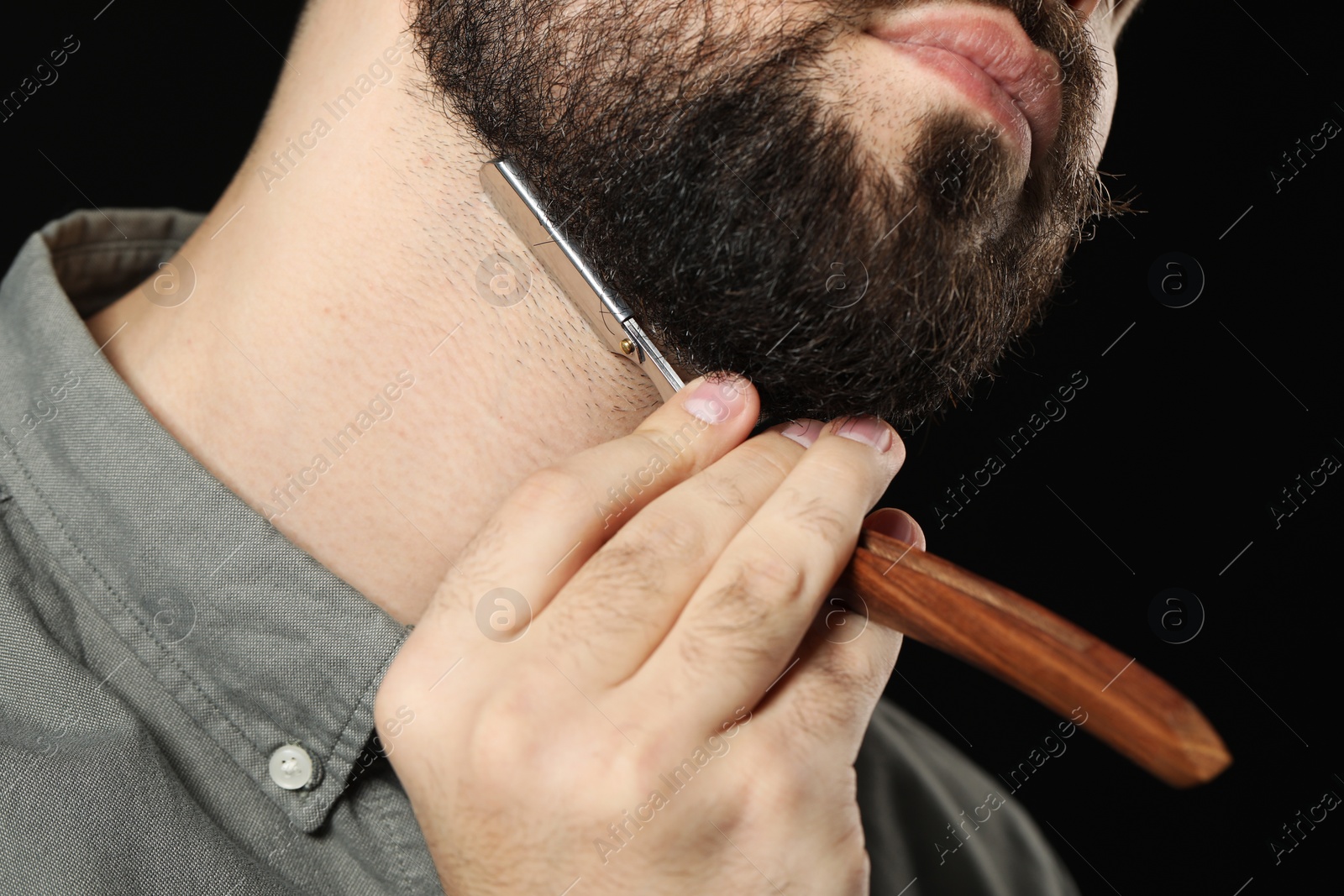 Photo of Handsome young man shaving beard with blade on black background, closeup
