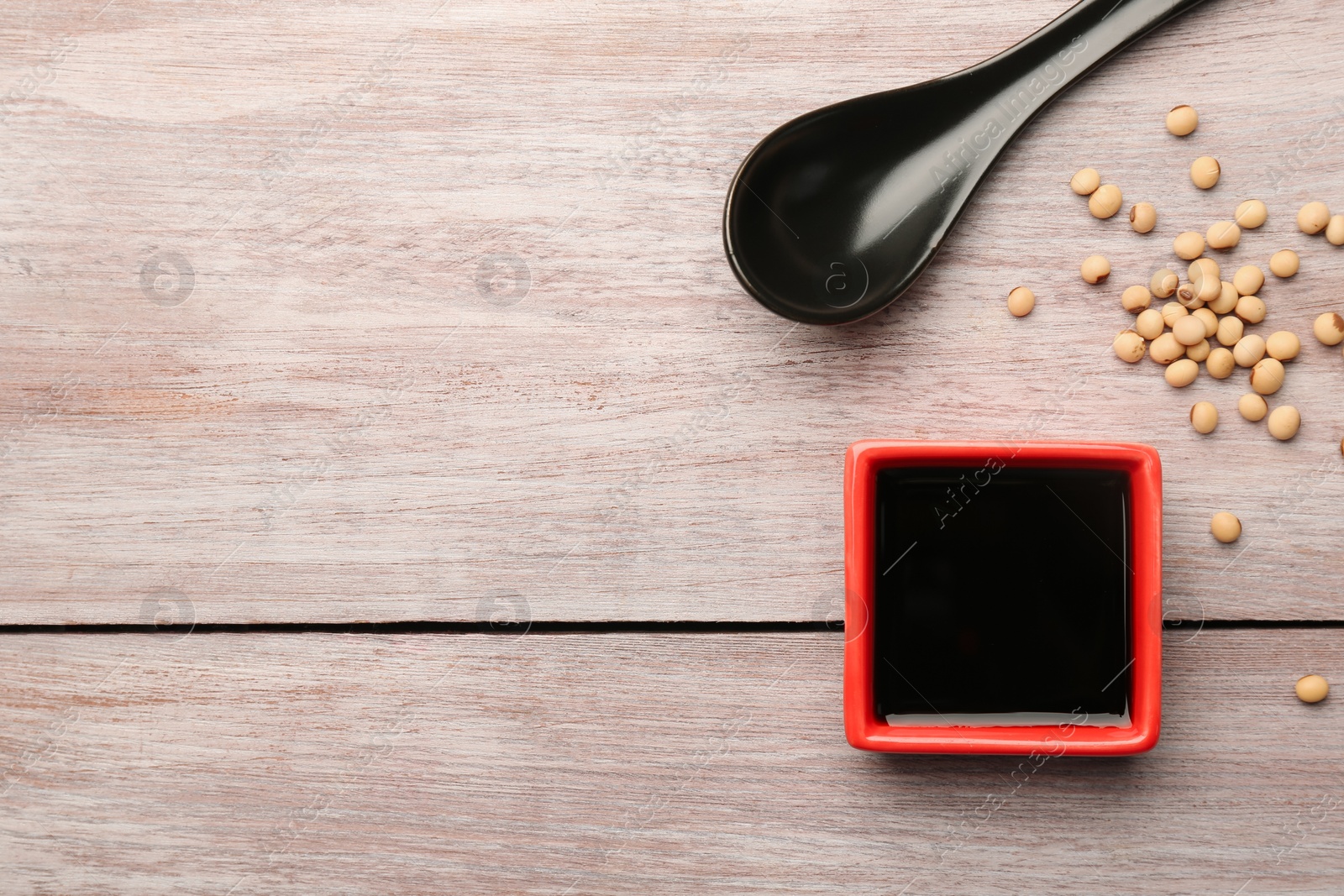 Photo of Soy sauce in bowl, soybeans and spoon on wooden table, flat lay. Space for text