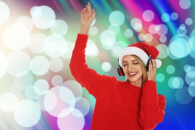 Happy woman in Santa hat listening to Christmas music with headphones on bright background, bokeh effect