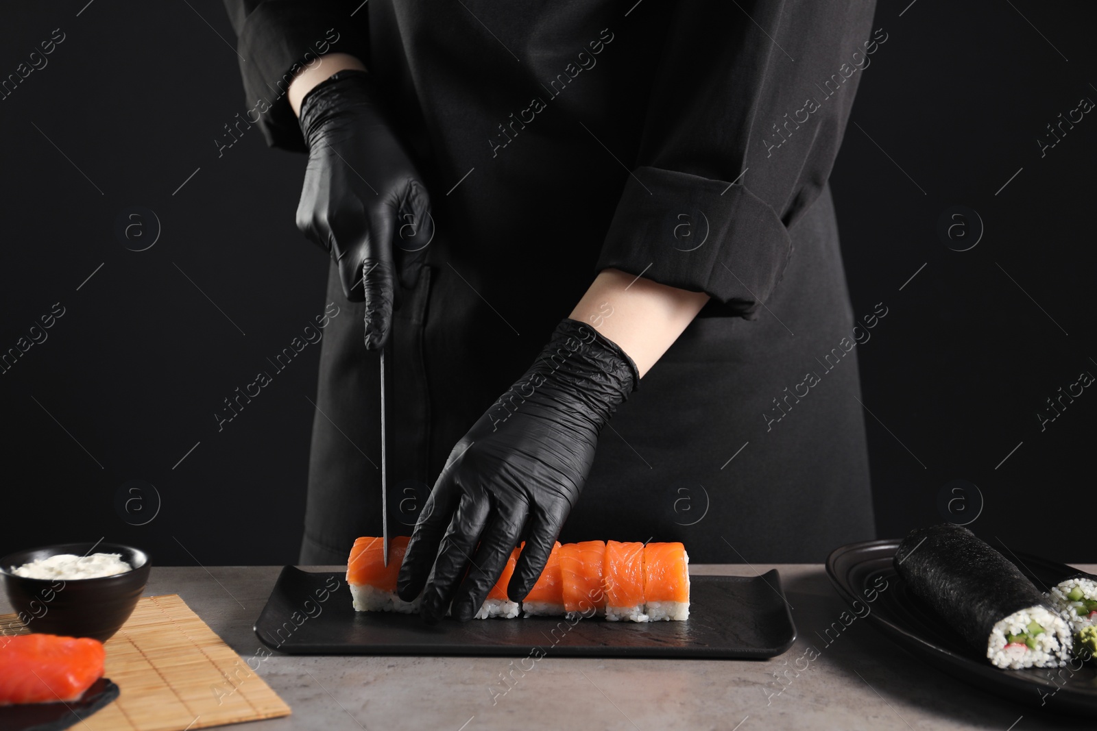 Photo of Chef in gloves cutting sushi roll at grey table, closeup