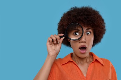 Surprised woman looking through magnifier glass on light blue background, space for text