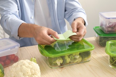Photo of Man sticking paper note onto container with fresh broccoli at wooden table, closeup. Food storage
