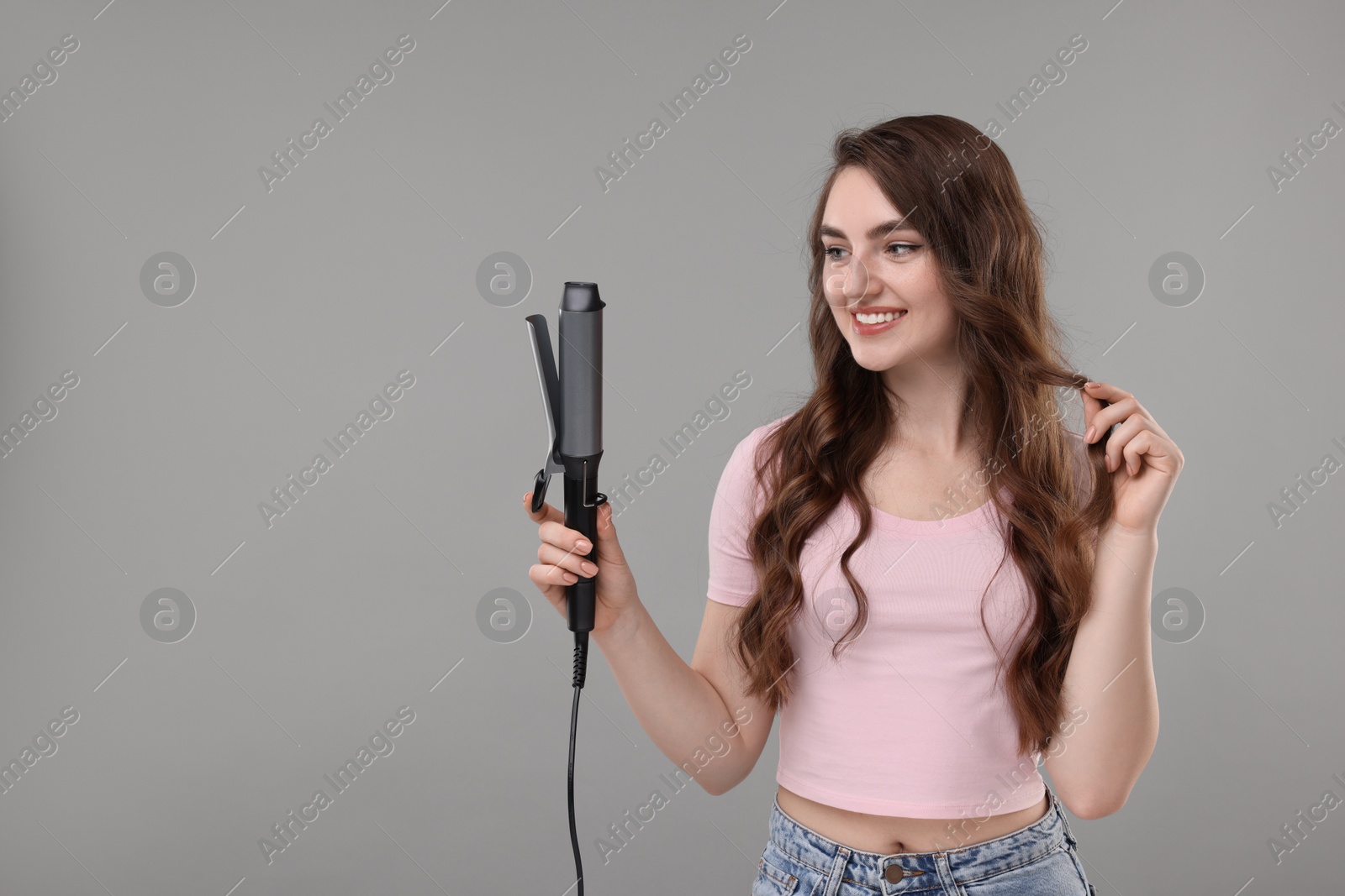 Photo of Happy young woman with beautiful hair holding curling iron on grey background, space for text