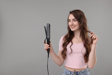 Happy young woman with beautiful hair holding curling iron on grey background, space for text