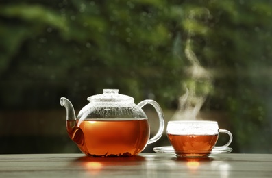 Photo of Teapot and cup of hot tea on wooden table against blurred background, space for text