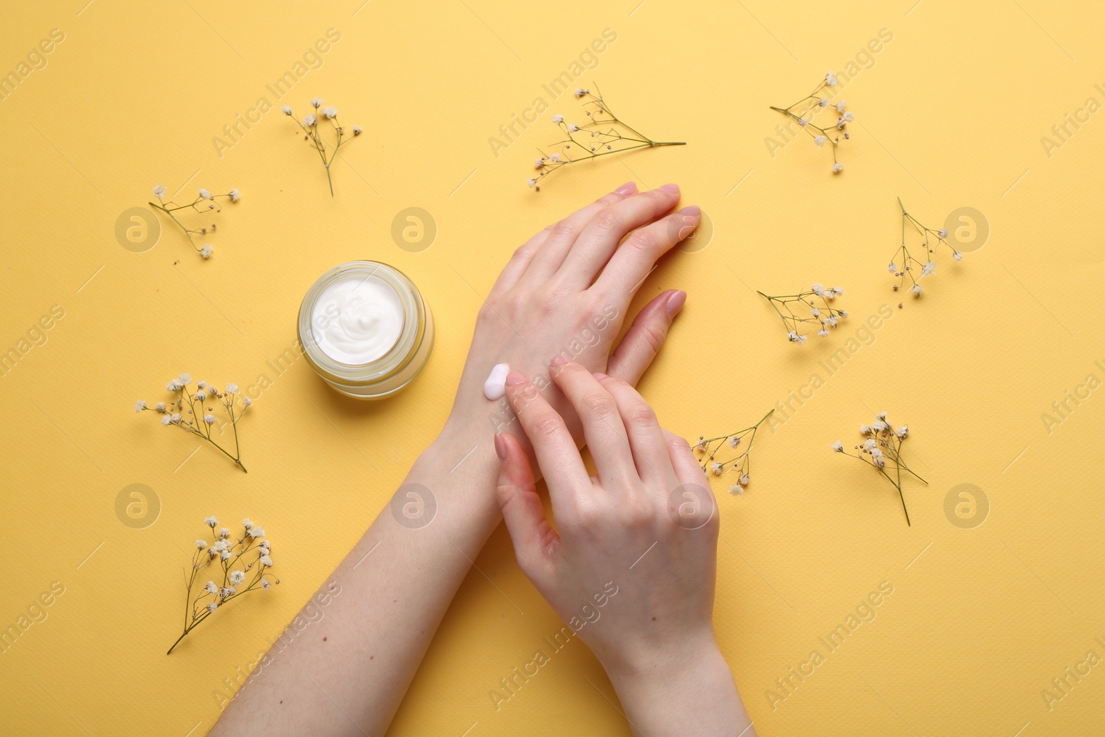 Photo of Woman applying hand cream and flowers on yellow background, top view