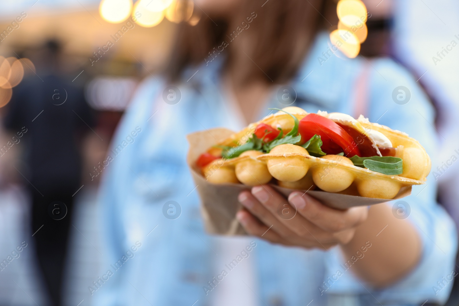 Photo of Young woman holding delicious bubble waffle with tomato and arugula outdoors, closeup