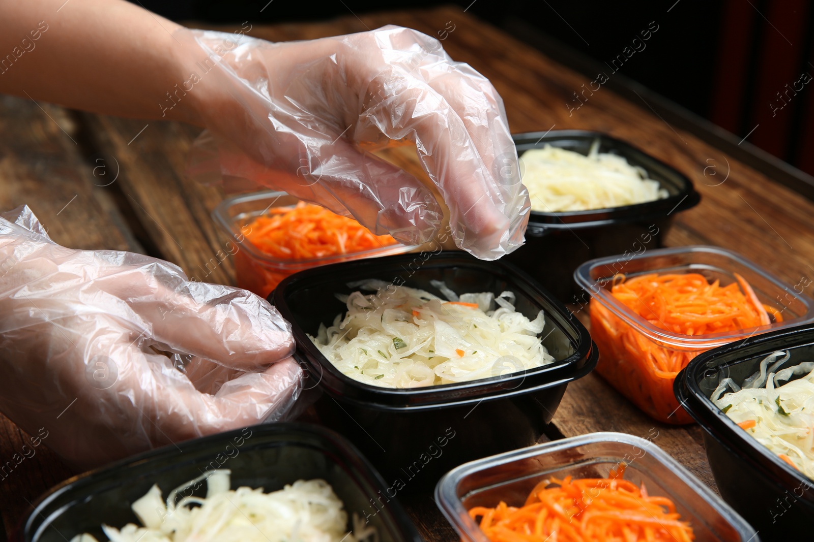 Photo of Waiter in gloves putting salads into containers at wooden table, closeup. Food delivery service