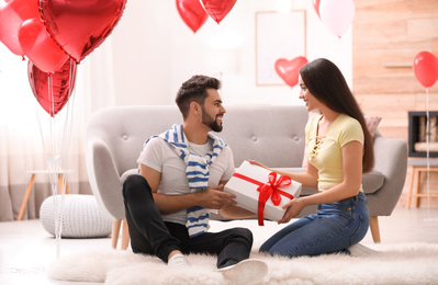Photo of Lovely young couple with gift box in living room decorated with heart shaped balloons. Valentine's day celebration